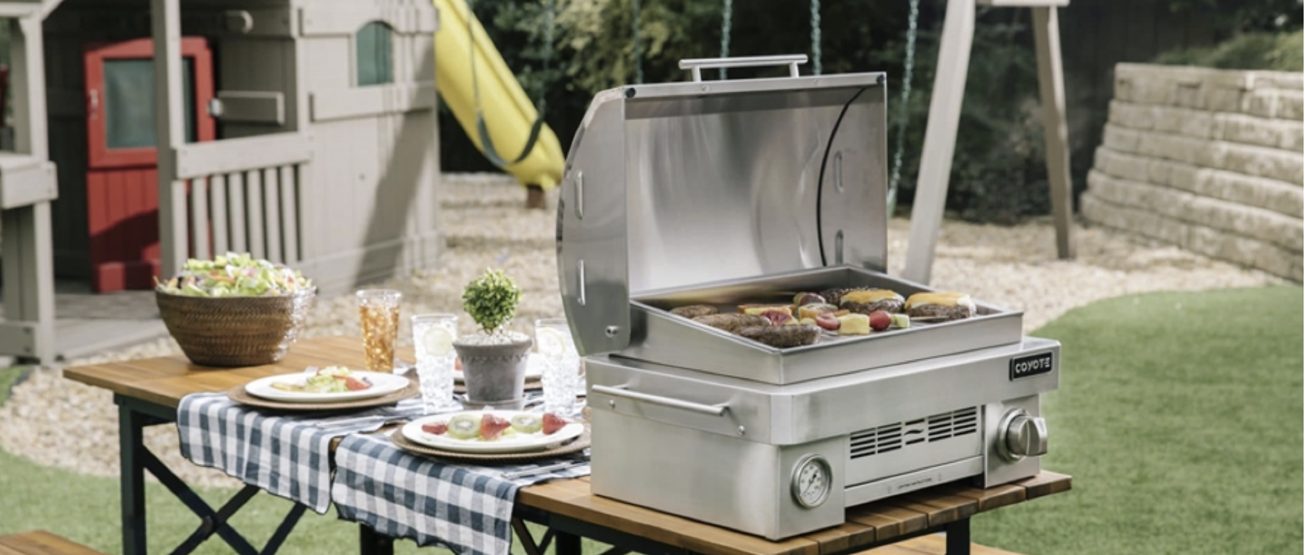 a portable grill on an outdoor picnic table with a swingset in the background